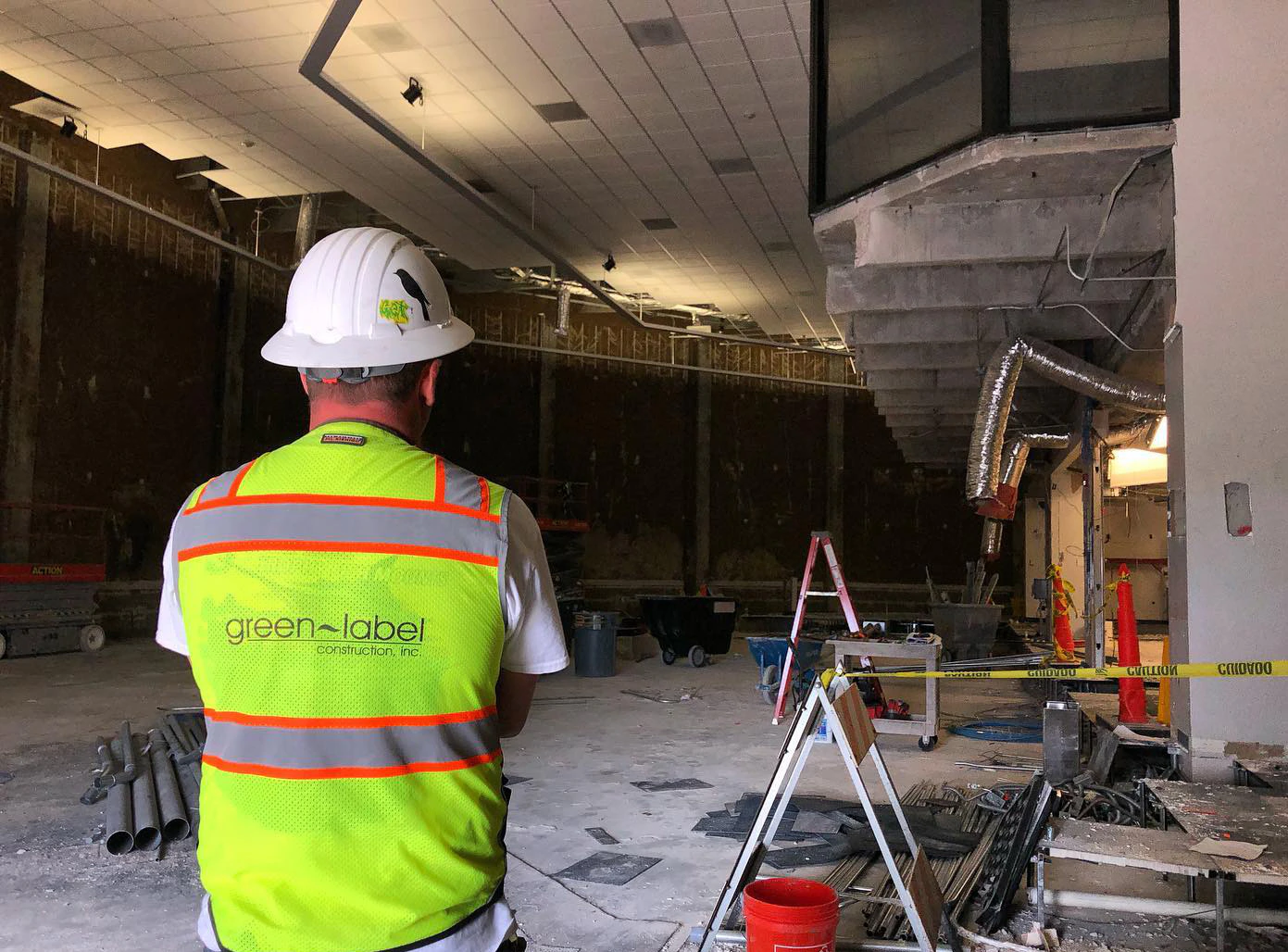 Construction worker viewed from behind wearing a neon yellow high-visibility vest with the Green Label Construction logo and a white hard hat, standing in an unfinished commercial interior under renovation with exposed ceilings, ducts, and construction materials