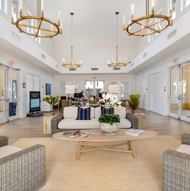 Bright double-height lobby featuring circular gold chandeliers, a central seating area with beige sofa and coffee table, potted orchid, and large windows looking out to outdoor spaces